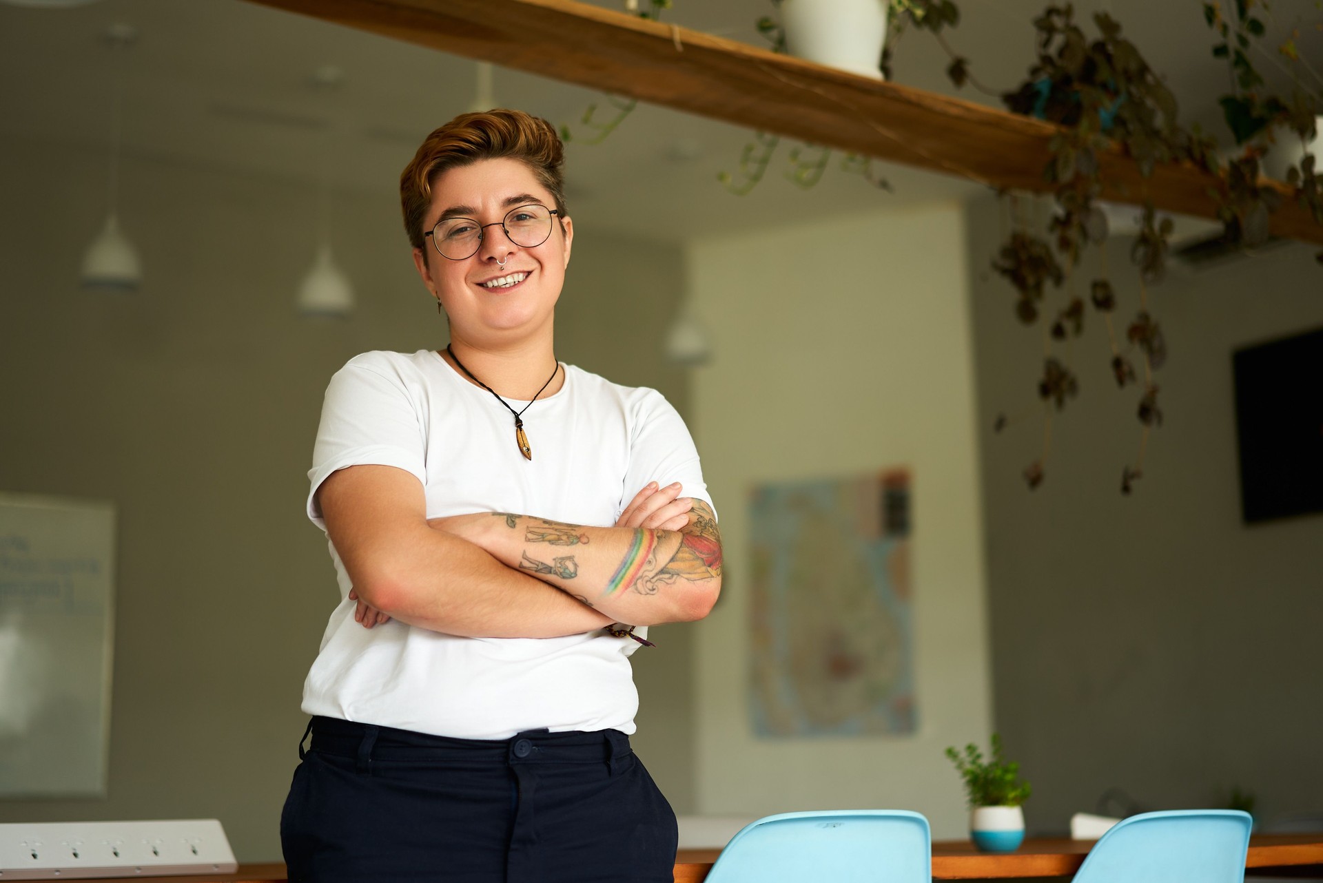 Confident Gen Z individual with tattoos stands in modern office. Transgender young adult smiles, arms crossed, wearing casual attire, representing workplace inclusion, gender identity pride.