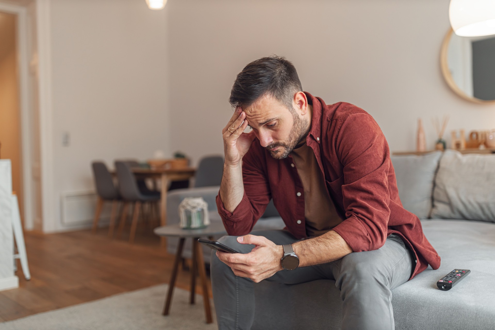 An anxious adult man reading bad news on his phone while sitting on a couch