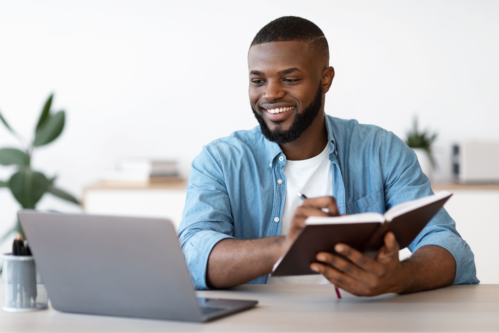 Black Millennial Freelancer Guy Taking Notes While Working On Laptop At Home